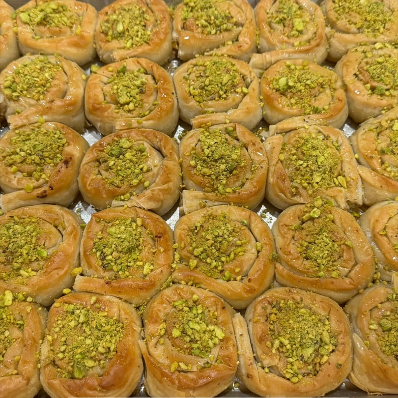 A tray of golden-brown, spiral-shaped baklava pastries, generously topped with crushed green pistachios.