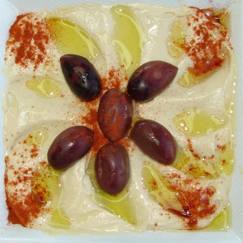 Hummus topped with whole olives, olive oil, and paprika, served in a white dish.